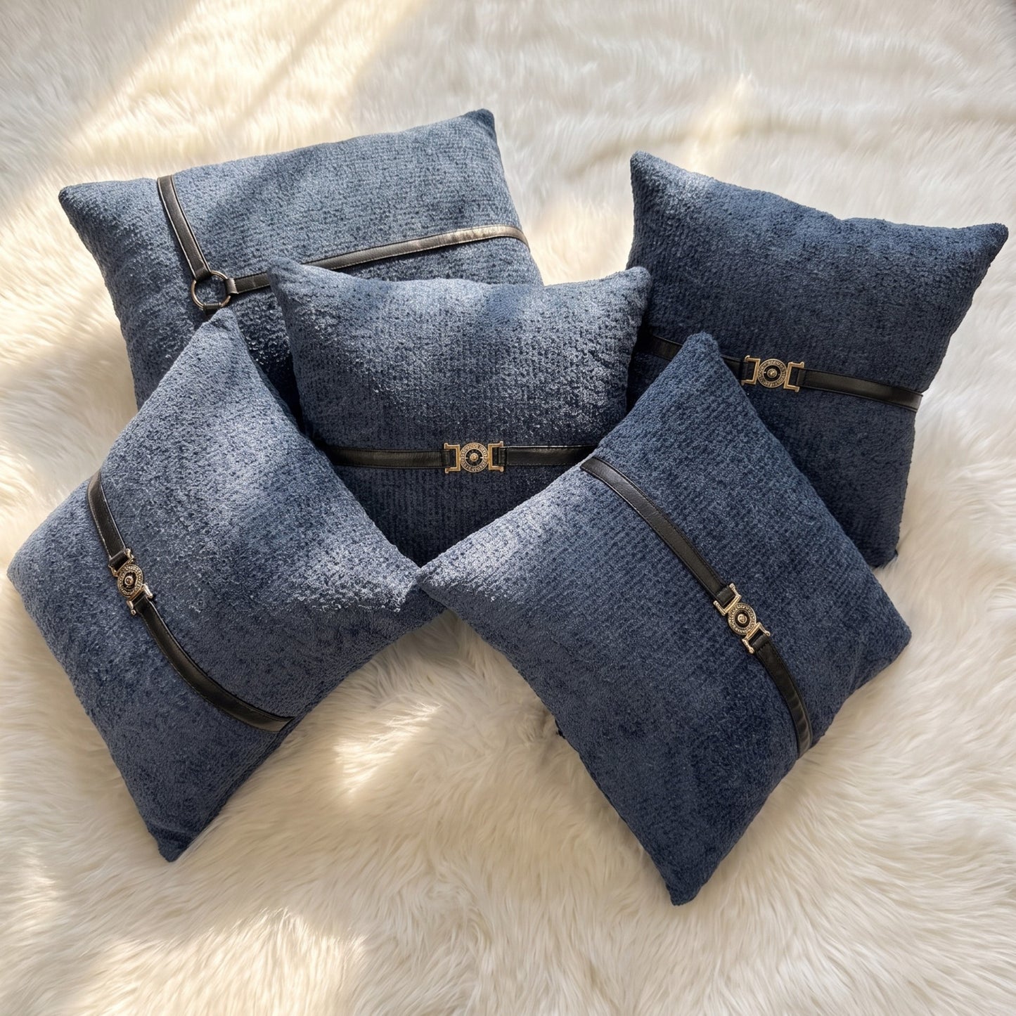 Five slate blue bouclé pillow covers arranged together, featuring black faux leather straps with gold-tone buckle and ring hardware.