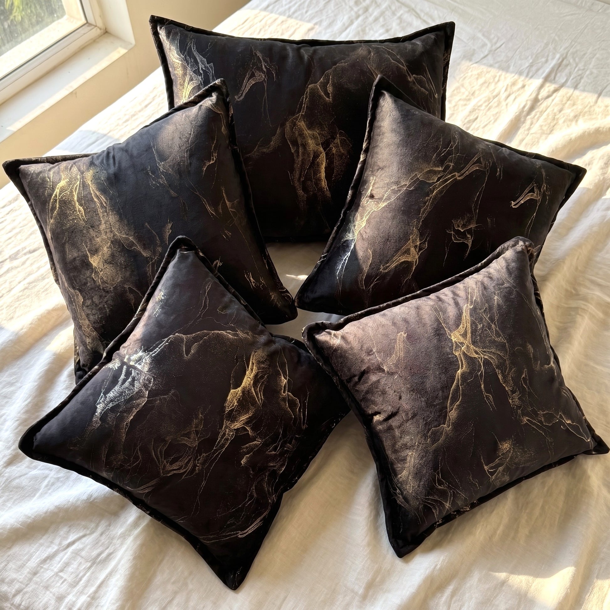Five black velvet pillow covers with flowing gold-toned abstract smoke print arranged together in natural window light.