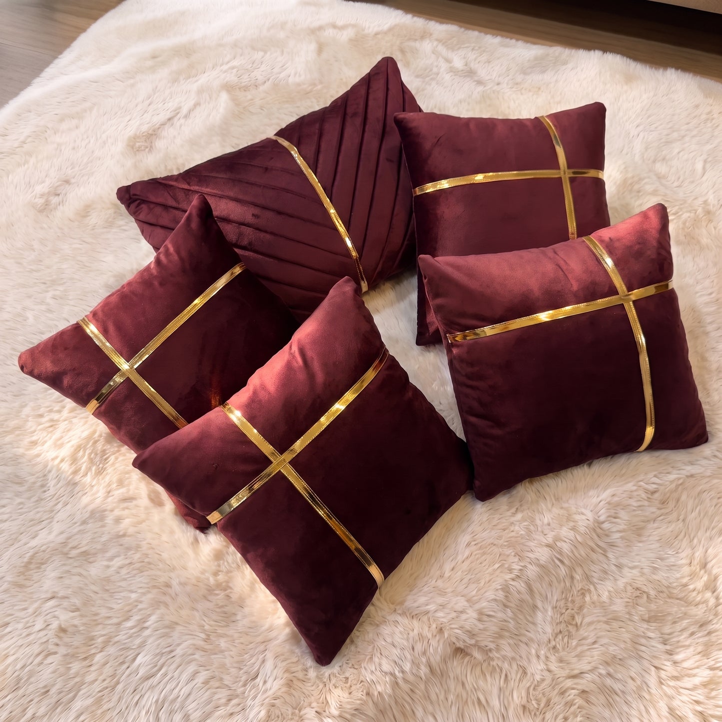 Five burgundy velvet pillow covers arranged together, featuring gold-tone trim details and one dimensional pleated rectangular lumbar cover.