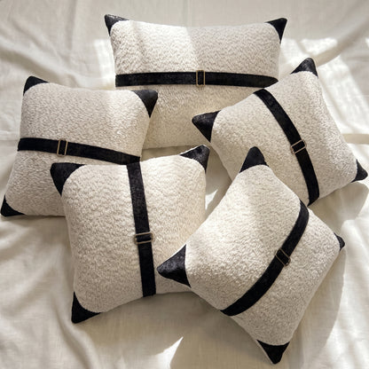 Five white bouclé pillow covers arranged together with contrasting black printed velvet accents and bronze-tone buckle details.