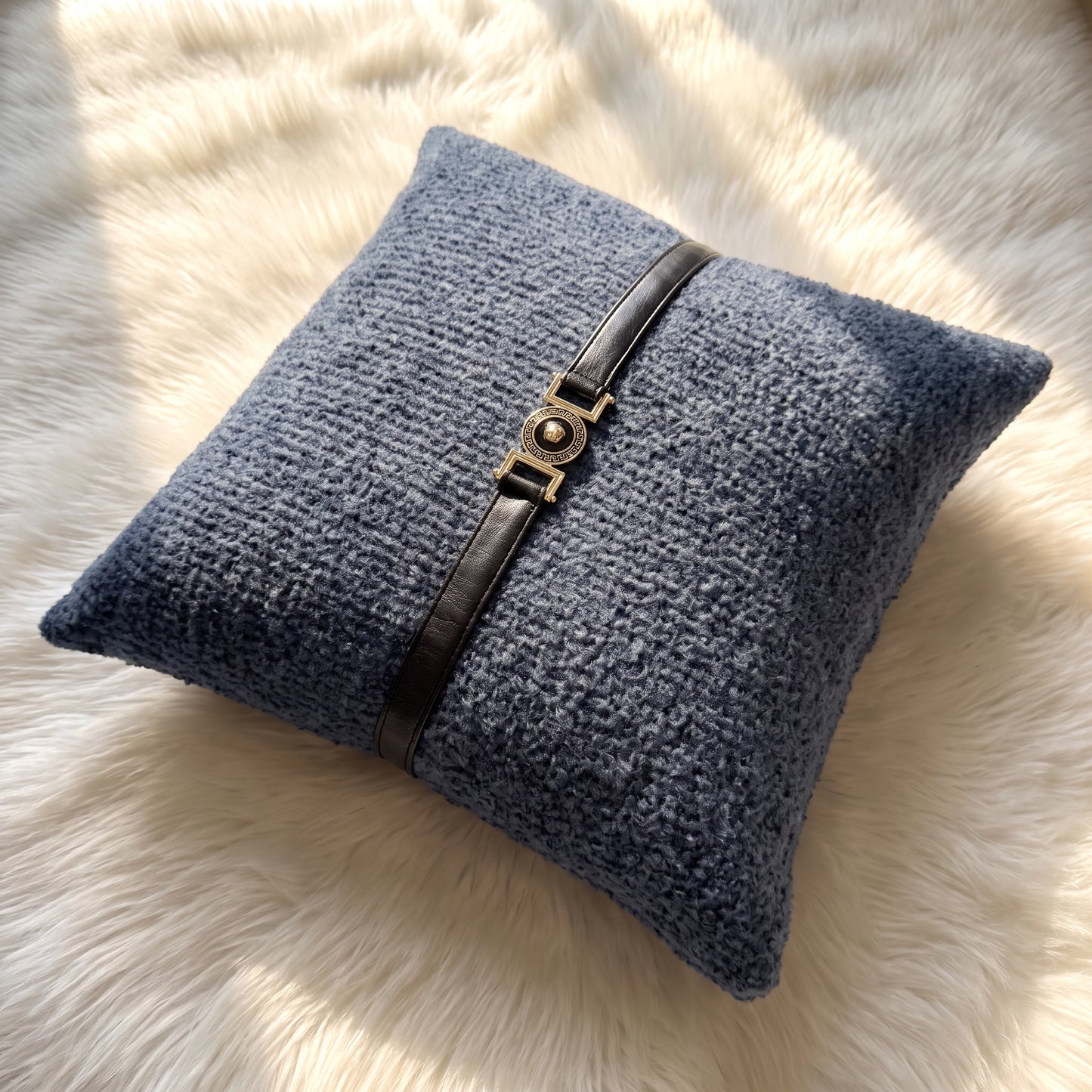 Slate blue bouclé square pillow cover with a black faux leather strap and decorative gold-tone circular buckle centered across the front.
