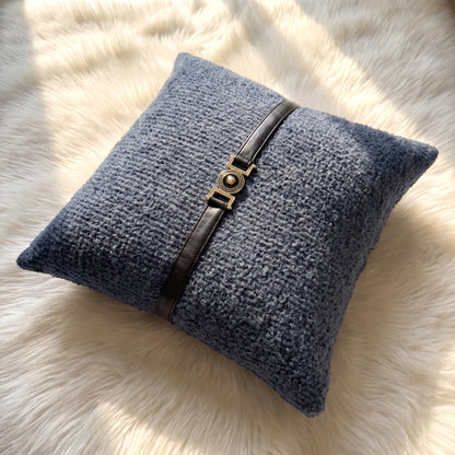 Slate blue bouclé square pillow cover with a black faux leather strap and decorative gold-tone circular buckle centered across the front.
