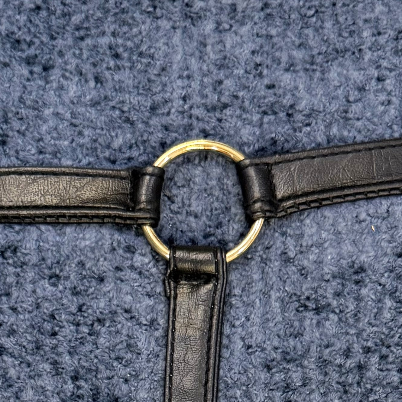 Close-up of black faux leather straps attached around a round gold-tone ring on textured slate blue bouclé fabric.
