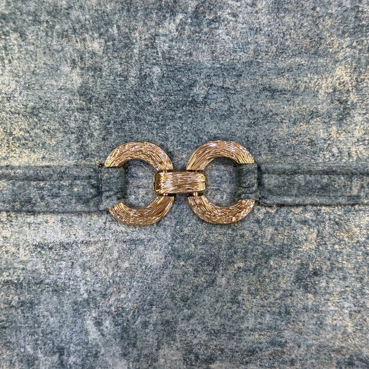Close-up of textured gold-tone decorative ring hardware attached to a blue-gray velvet strap.
