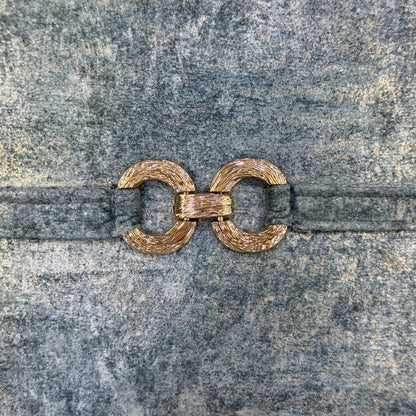 Close-up of textured gold-tone decorative ring hardware attached to a blue-gray velvet strap.
