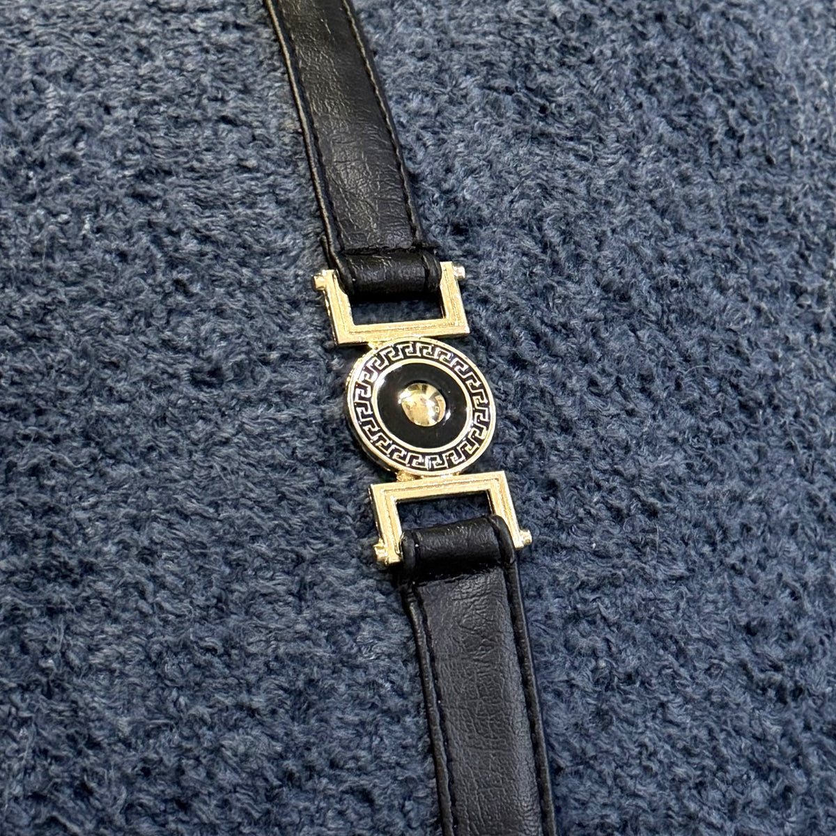 Close-up of gold-tone and black decorative buckle hardware attached to a black faux leather strap over slate blue bouclé.

