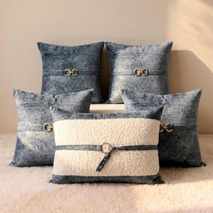 Four muted blue-gray velvet square pillow covers arranged around an ivory bouclé lumbar cover with gold-tone decorative hardware.